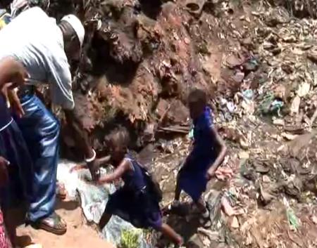 School children are helped across a stream as they move through a pile of garbage.
